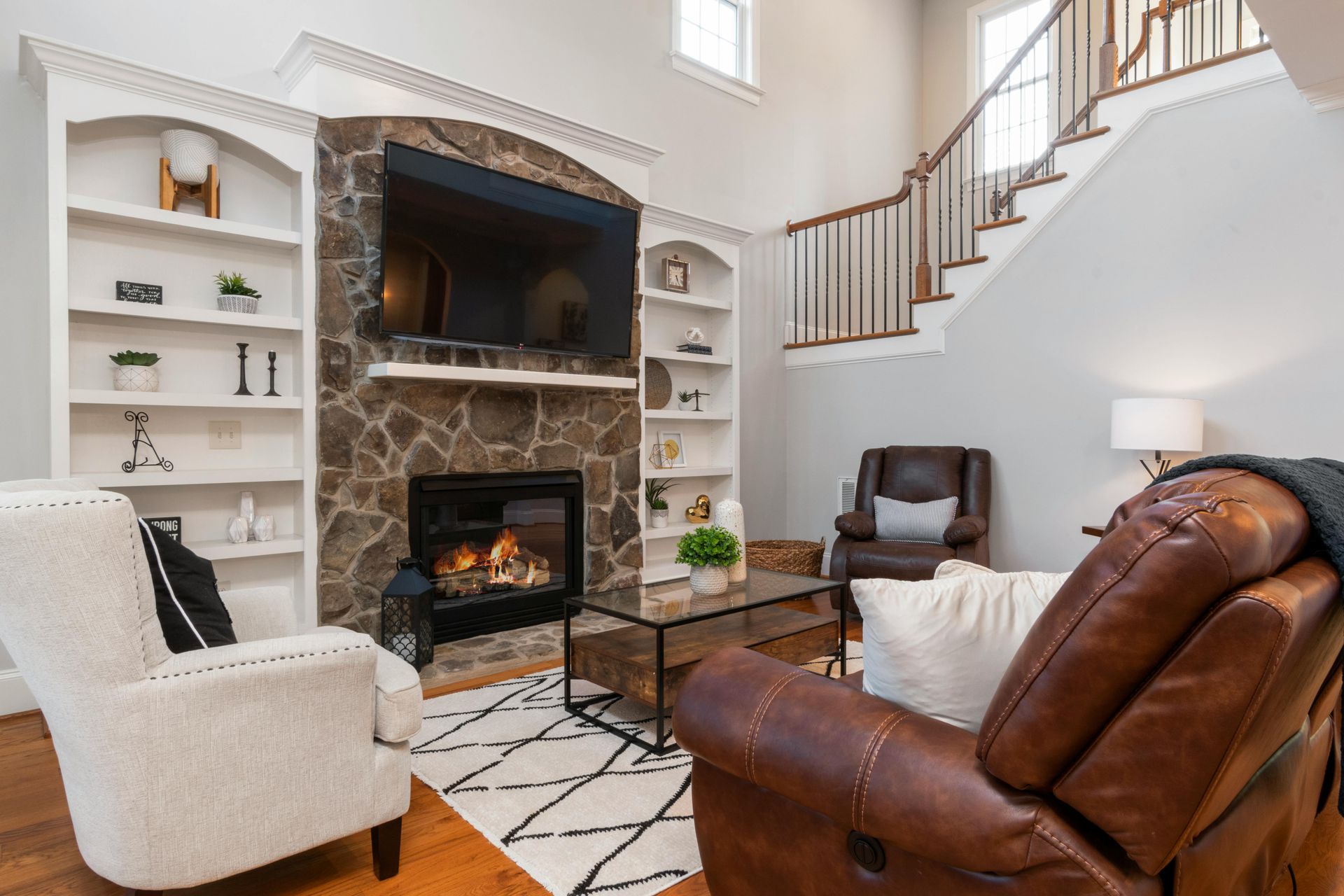 A living room with a stone fireplace, TV, white built-in shelves, stairs, and brown and white chairs on a patterned rug.