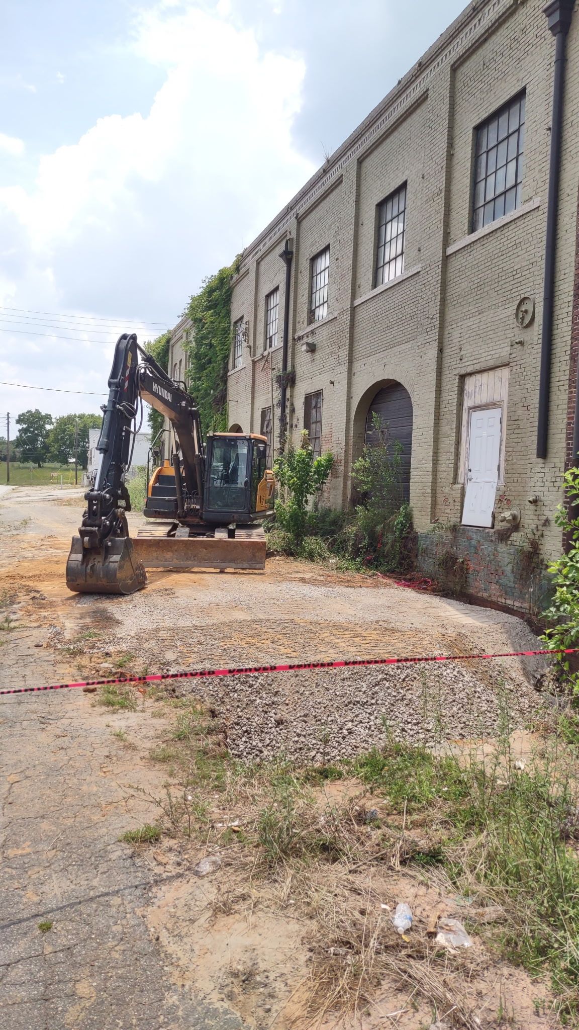 Excavator next to a brick building; gravel and dirt foreground, blue sky, red line.