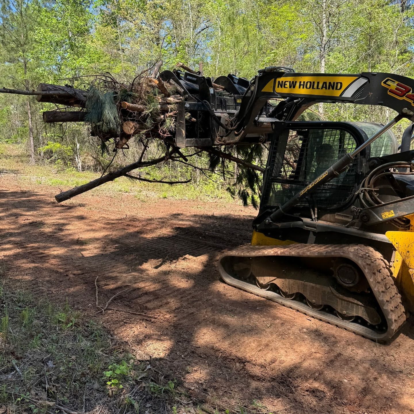 Yellow New Holland skid steer with a grapple lifting tree debris in a wooded area.