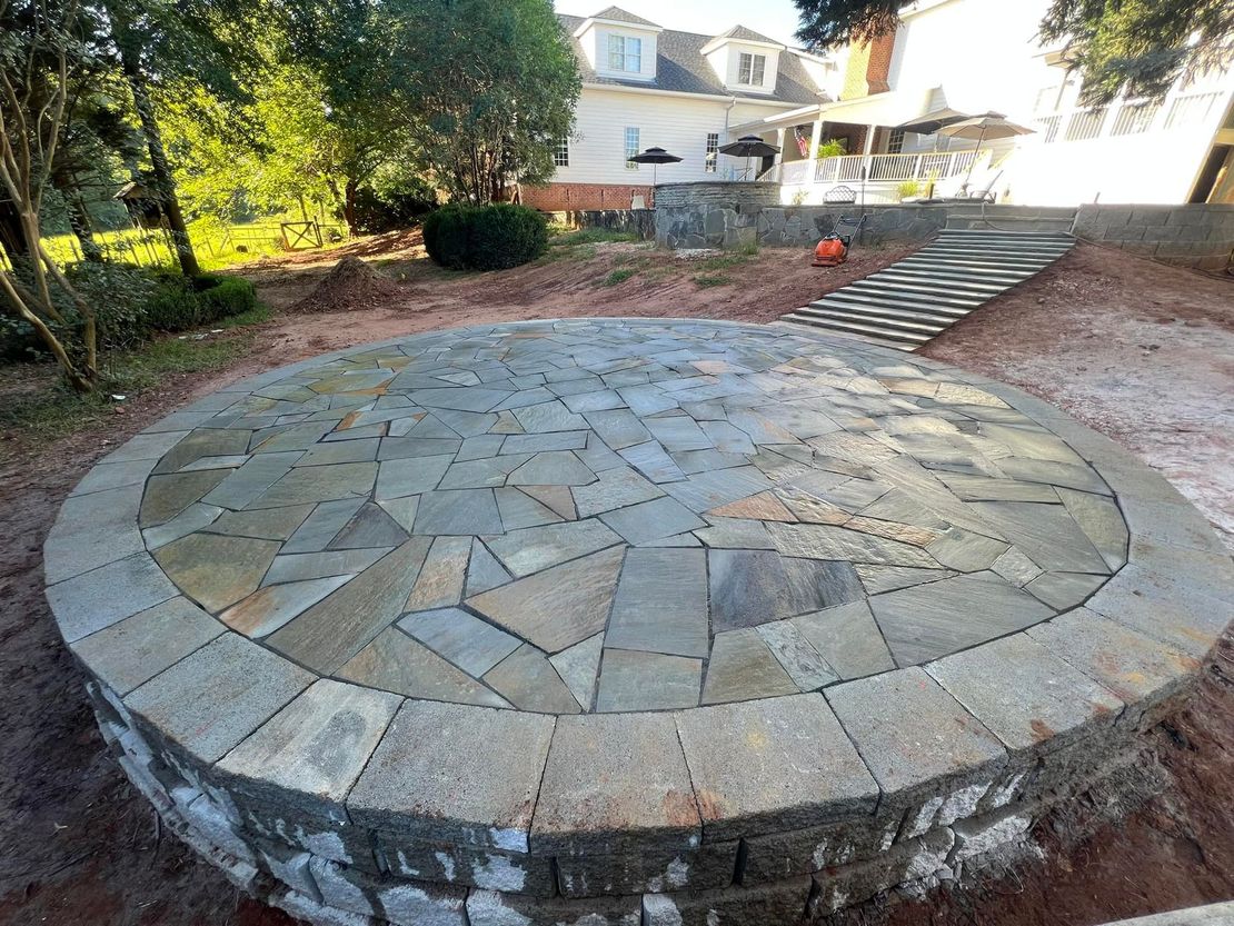 Circular patio built with stone, leading to stairs, in a backyard.