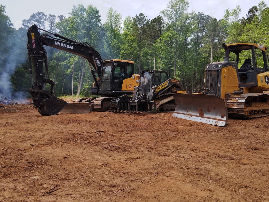 Heavy machinery clearing land: excavator, bulldozer, brown soil, trees in background.