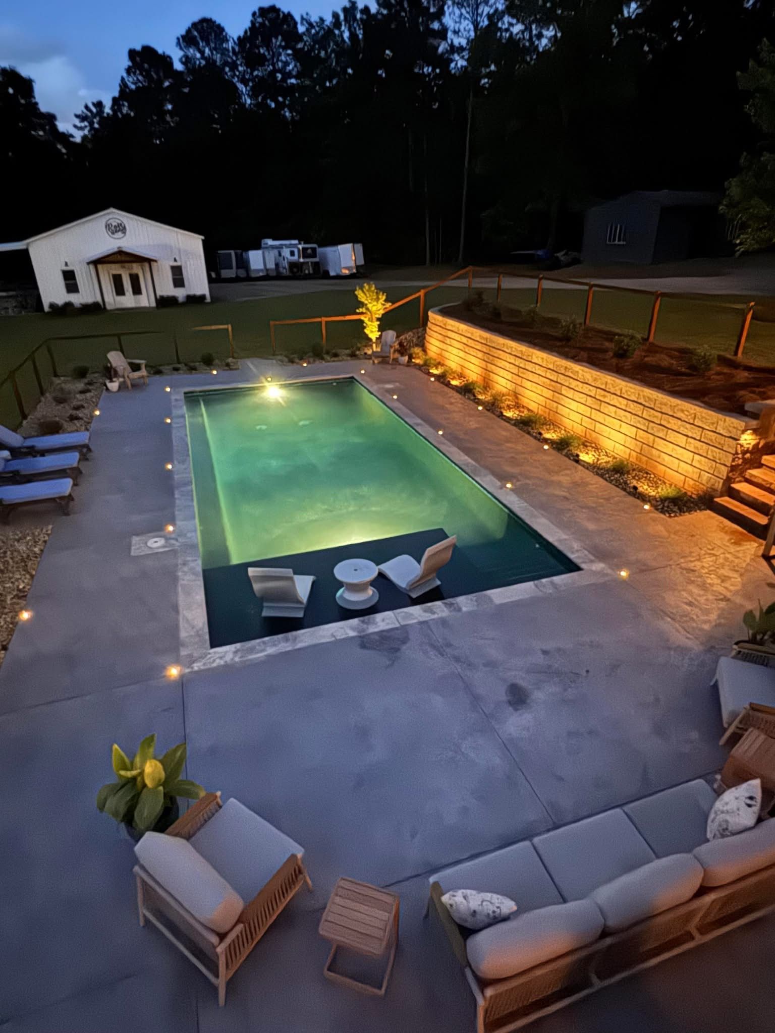 Night view of a lit pool surrounded by outdoor furniture and a garden wall, in a backyard.
