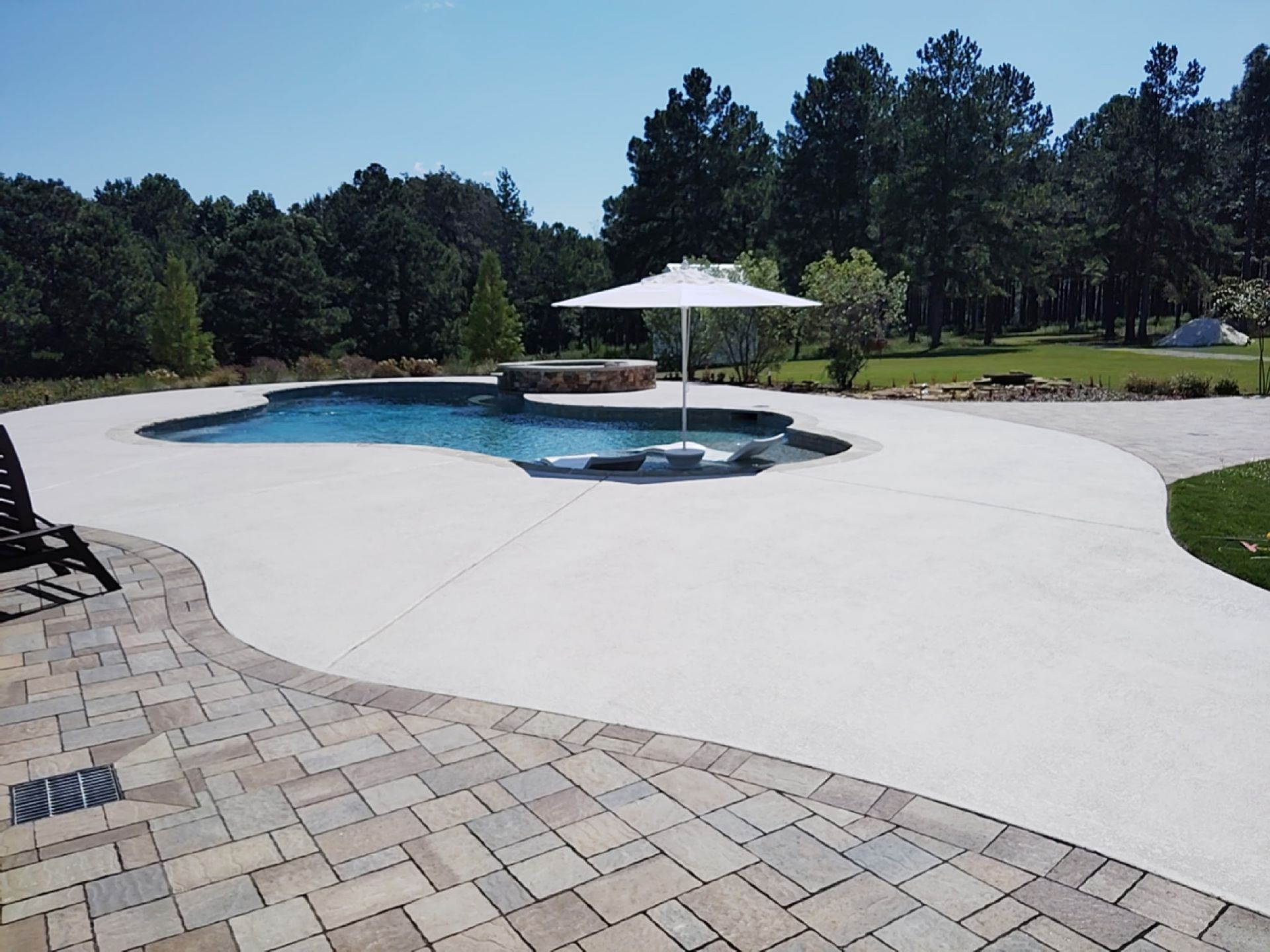 Pool with white umbrella, stone patio, and green trees under a clear blue sky.