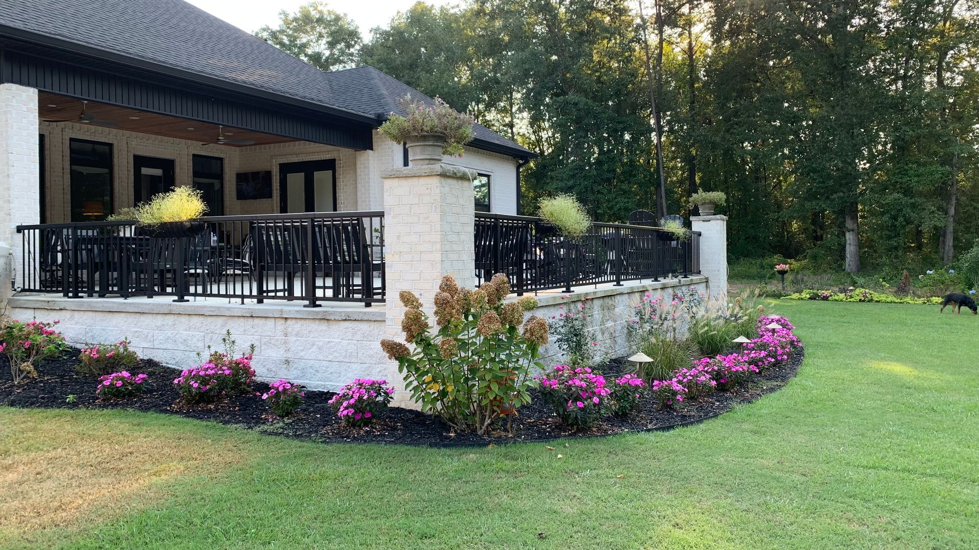 A house with a curved stone patio lined with pink flowers and black railing overlooking a green lawn.