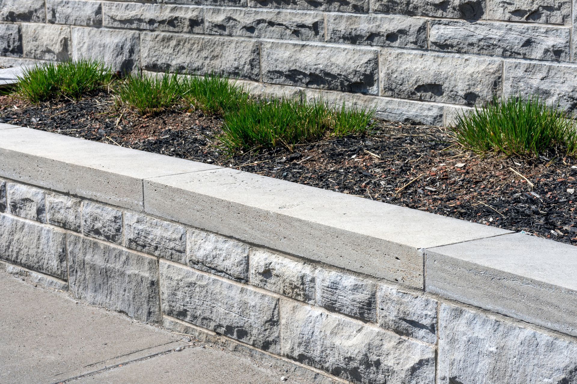Stone retaining wall with stepped landscaping and small green plants in front of a paved walkway Stone retaining wall with stepped landscaping and small green plants in front of a paved walkway