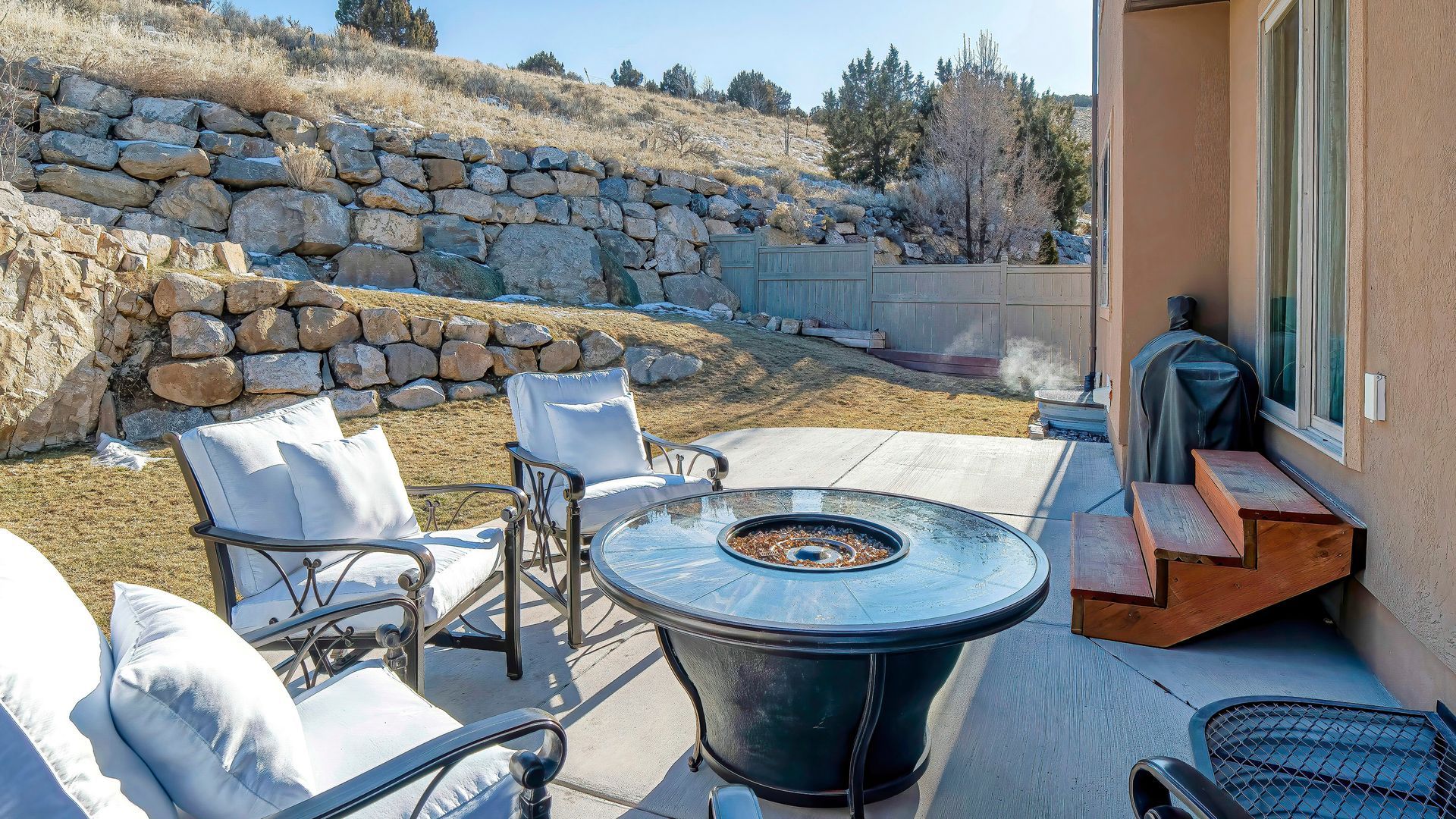 Sunlit stone patio with chairs, a fire pit table, and steps beside a house, backed by a rock retaining wall Sunlit stone patio with chairs, a fire pit table, and steps beside a house, backed by a rock retaining wall