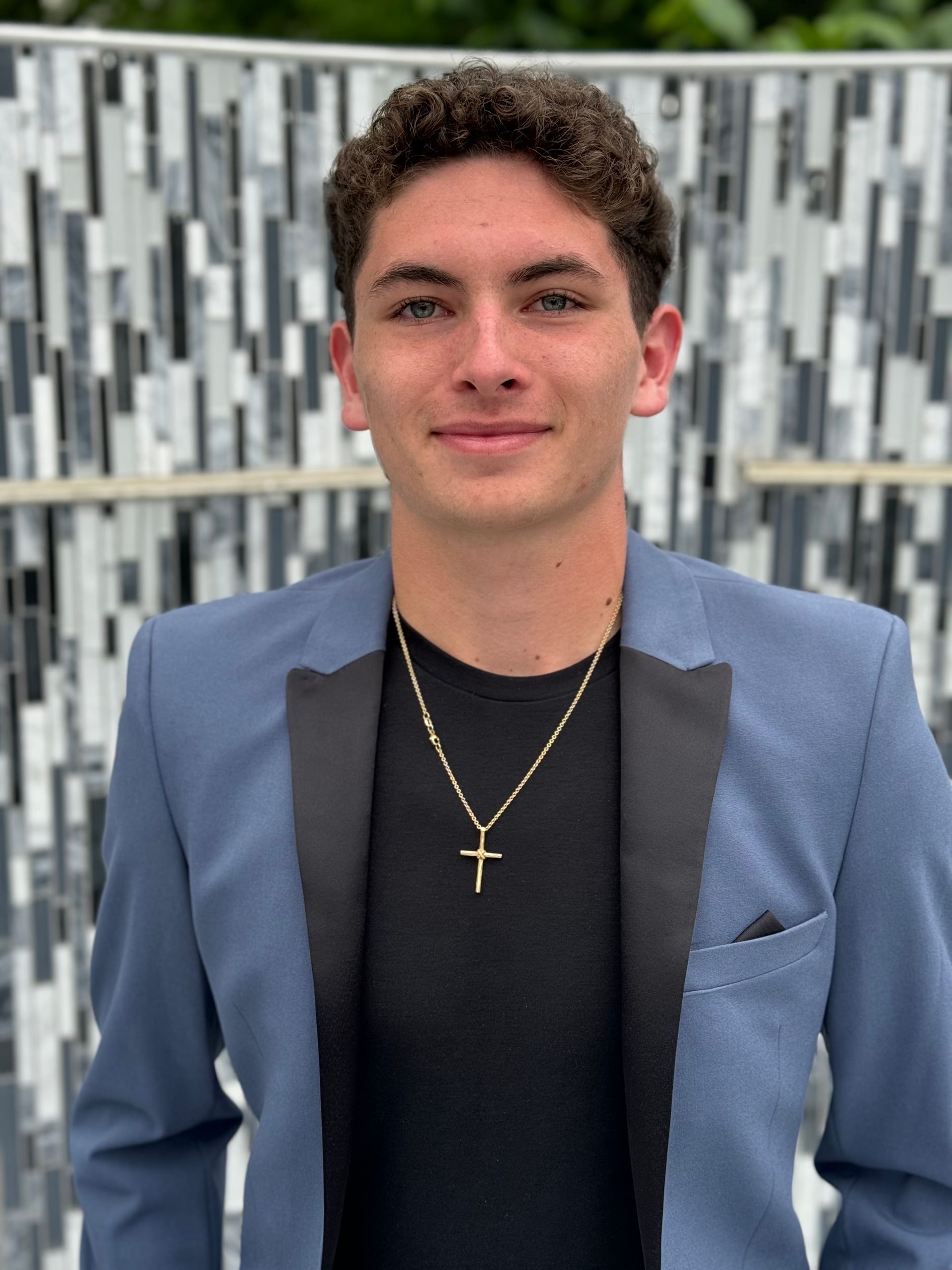 Young person in blue blazer and gold cross necklace, smiling, against a decorative wall.