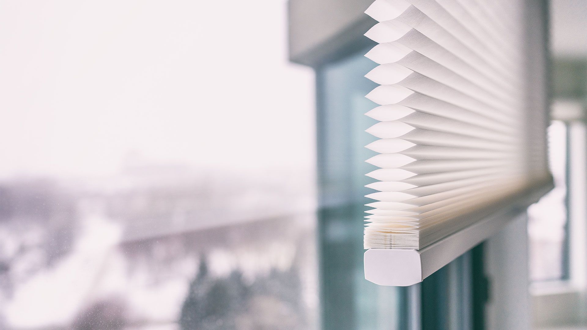 White pleated window blinds partially cover a window, snowy outdoor scene in background.
