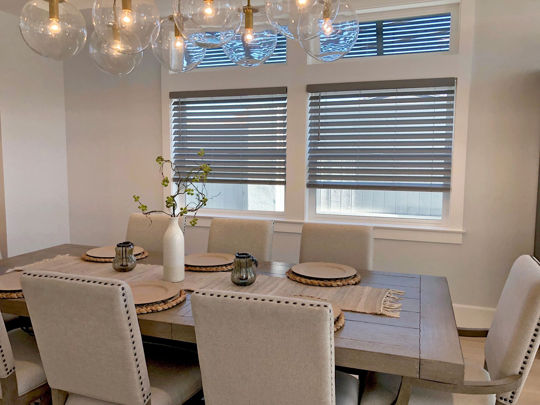 Dining room with a table set for a meal. Gray blinds cover the windows. Chandelier above the table.