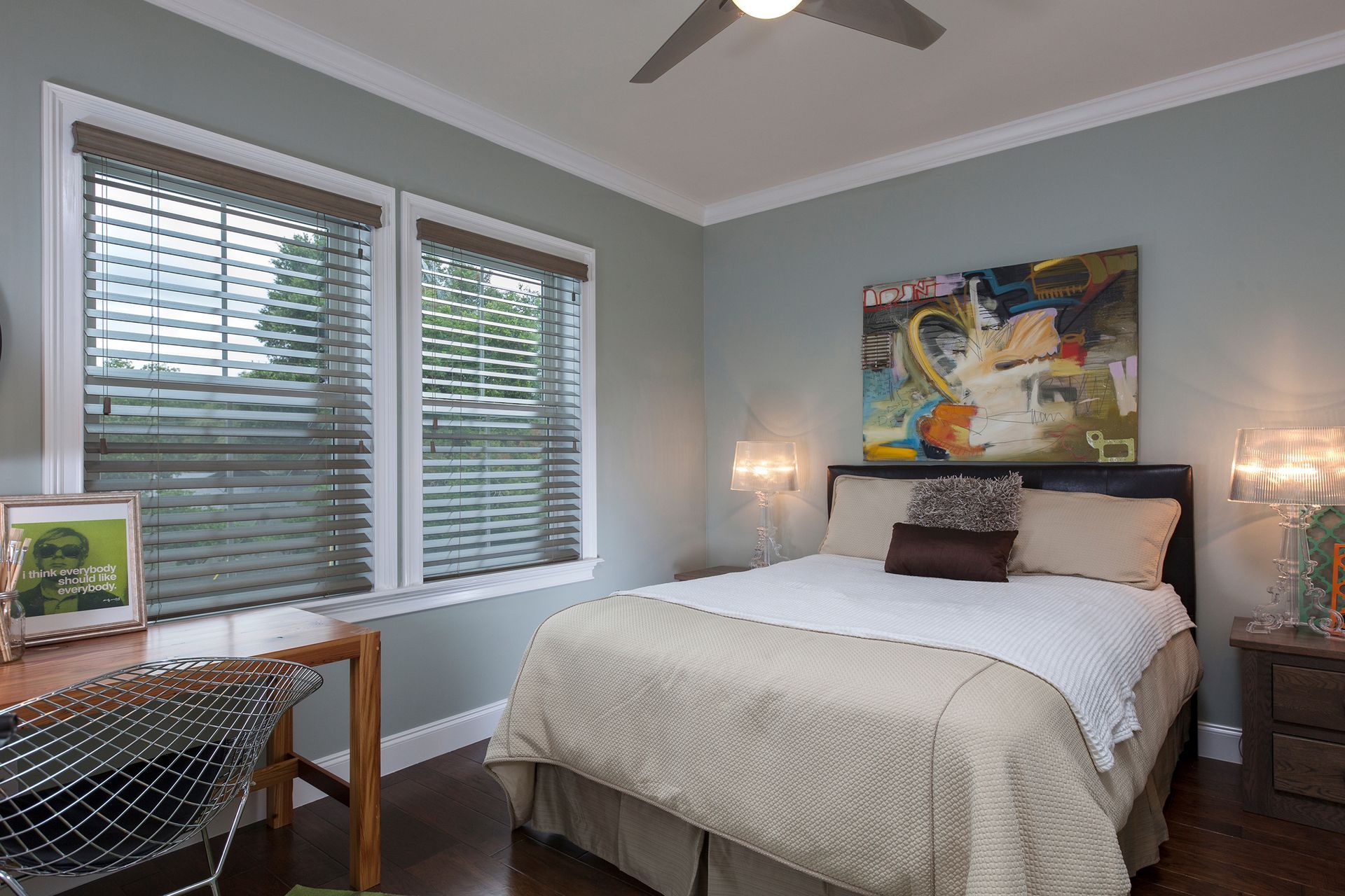 Bedroom with blue-green walls, a bed, desk by window with blinds. Abstract artwork hangs above the bed.