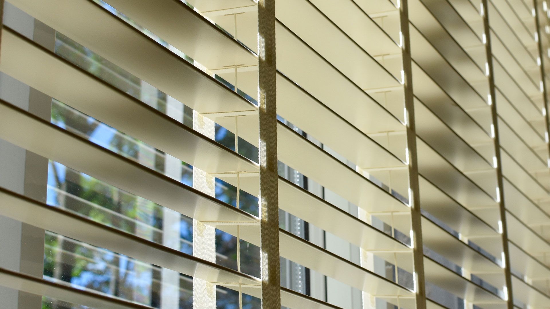 Close-up of beige window blinds, partially open, with a glimpse of trees and sky visible through the slats.