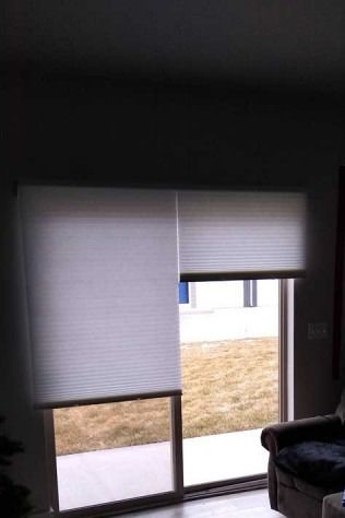 Two white pleated window blinds partially cover a glass door, revealing a yard and chair inside.