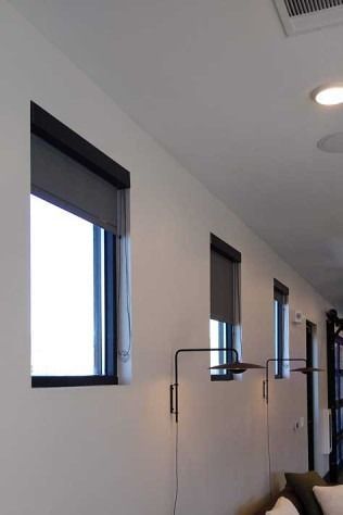 Three windows with dark roller shades in a white-walled room, two wall lamps below them.