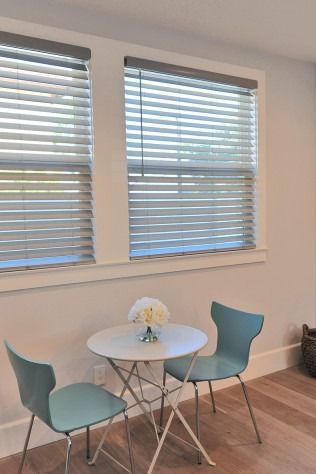 A small table with two teal chairs sits in front of a window with blinds, lit by natural light.