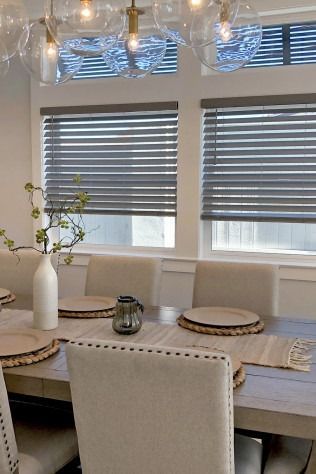 Dining room with a long wooden table, neutral chairs, and sheer blinds on windows.