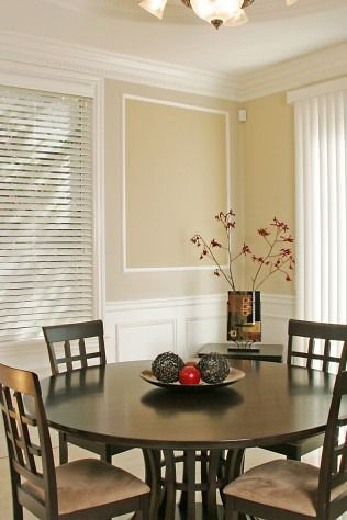 Dining room with round dark table, chairs, and decorative vase. Light walls, window blinds.