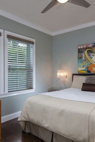 Bedroom with a bed, window with blinds, and artwork on a light blue wall. A ceiling fan is visible.