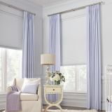 A bright nursery with pale blue curtains, blinds, an armchair, and a bedside table with a lamp and flowers.