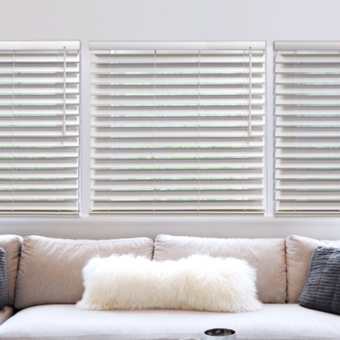 Three white blinds over a light-colored couch with decorative pillows.