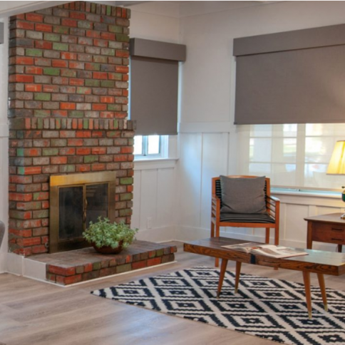 Cozy living room with brick fireplace, gray blinds, wood furniture, and geometric rug.