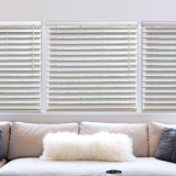 Cream-colored blinds over a white sectional sofa with fluffy and gray pillows.