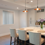 Kitchen with island, bar stools, and pendant lights. Window with vertical blinds. White cabinets and a vase of flowers.