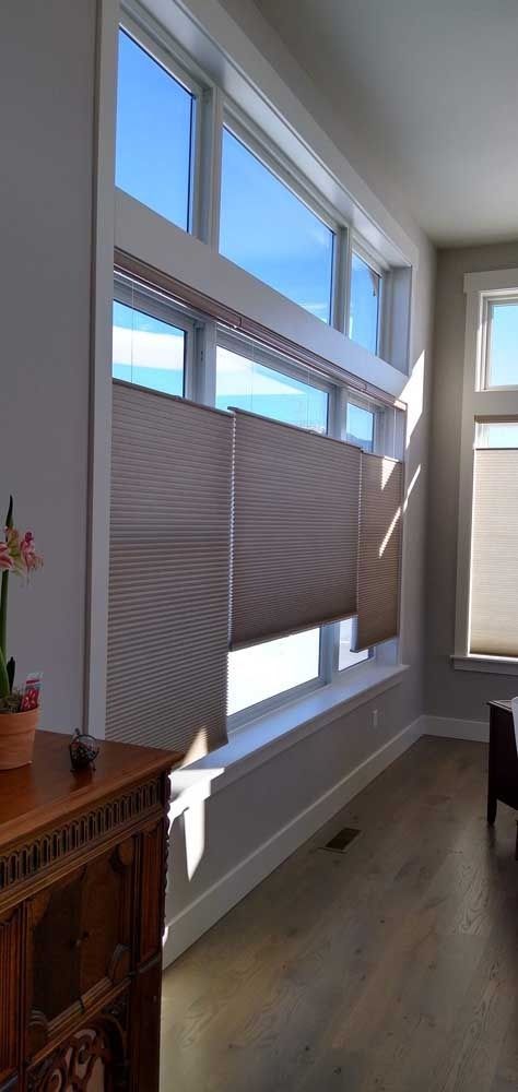 Large windows with cellular shades in a bright room. Sunlight streams in, illuminating the wooden floor.