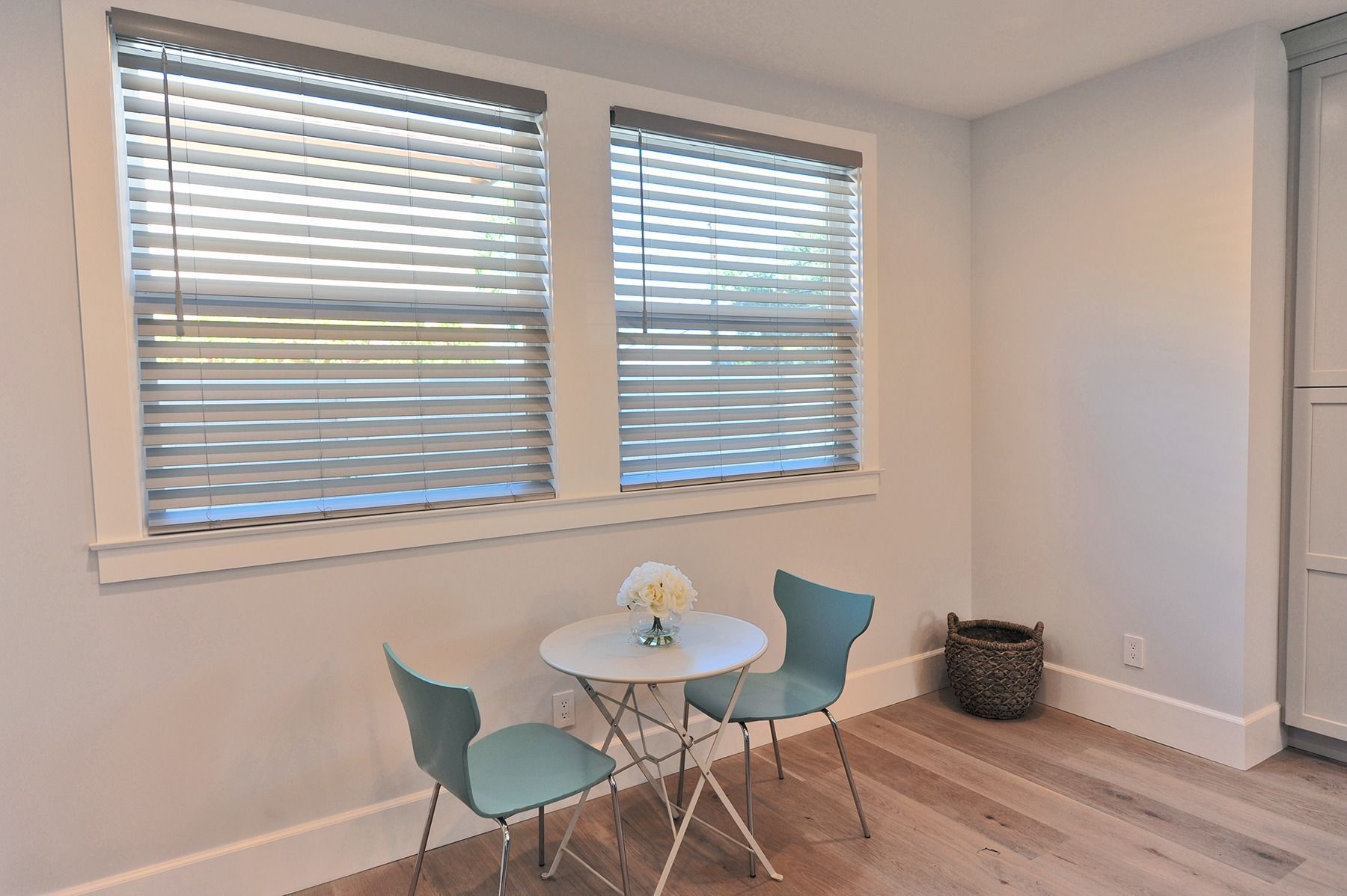 Small table and two teal chairs by a window with blinds, in a bright room with light wood floors.