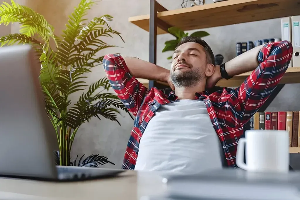 A man is sitting at a desk with his hands behind his head in front of a laptop computer.