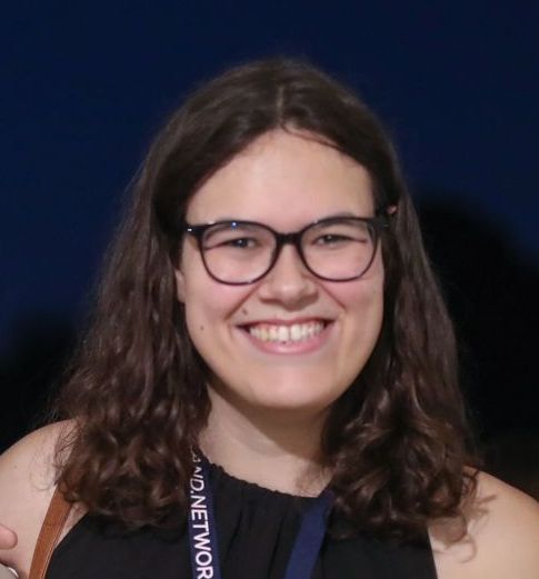A smiling woman with shoulder length, dark brown hair, wearing black, thick-rimmed spectacles and a black dress.