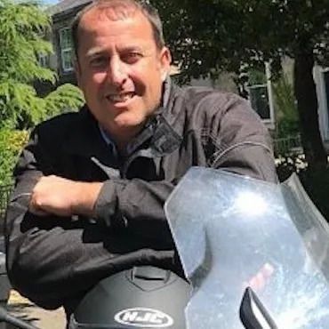 A smiling man, wearing a black button up shirt, sits with his arms crossed.