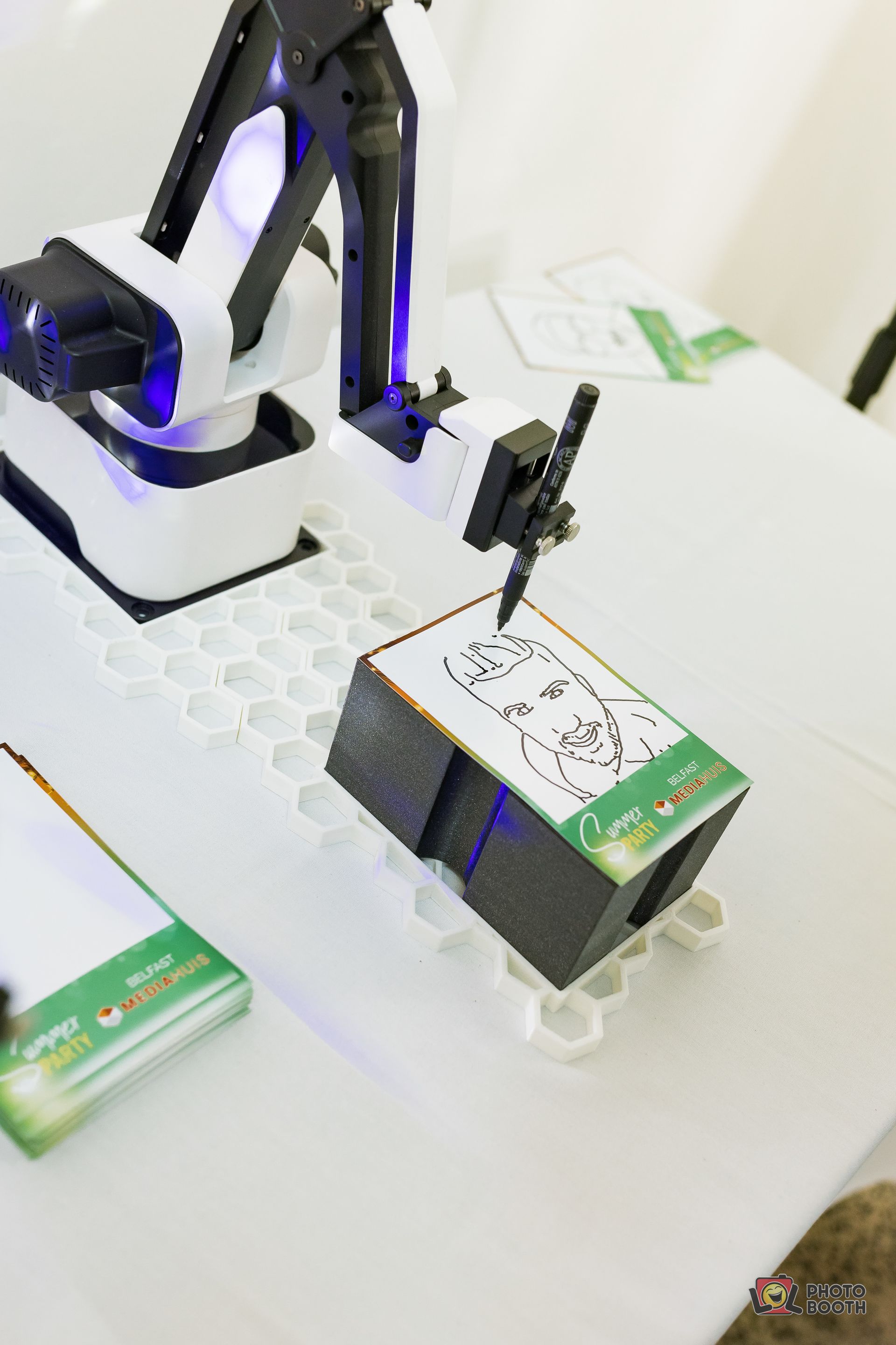 Robotic arm drawing a portrait on a small, stackable platform, on a white table.