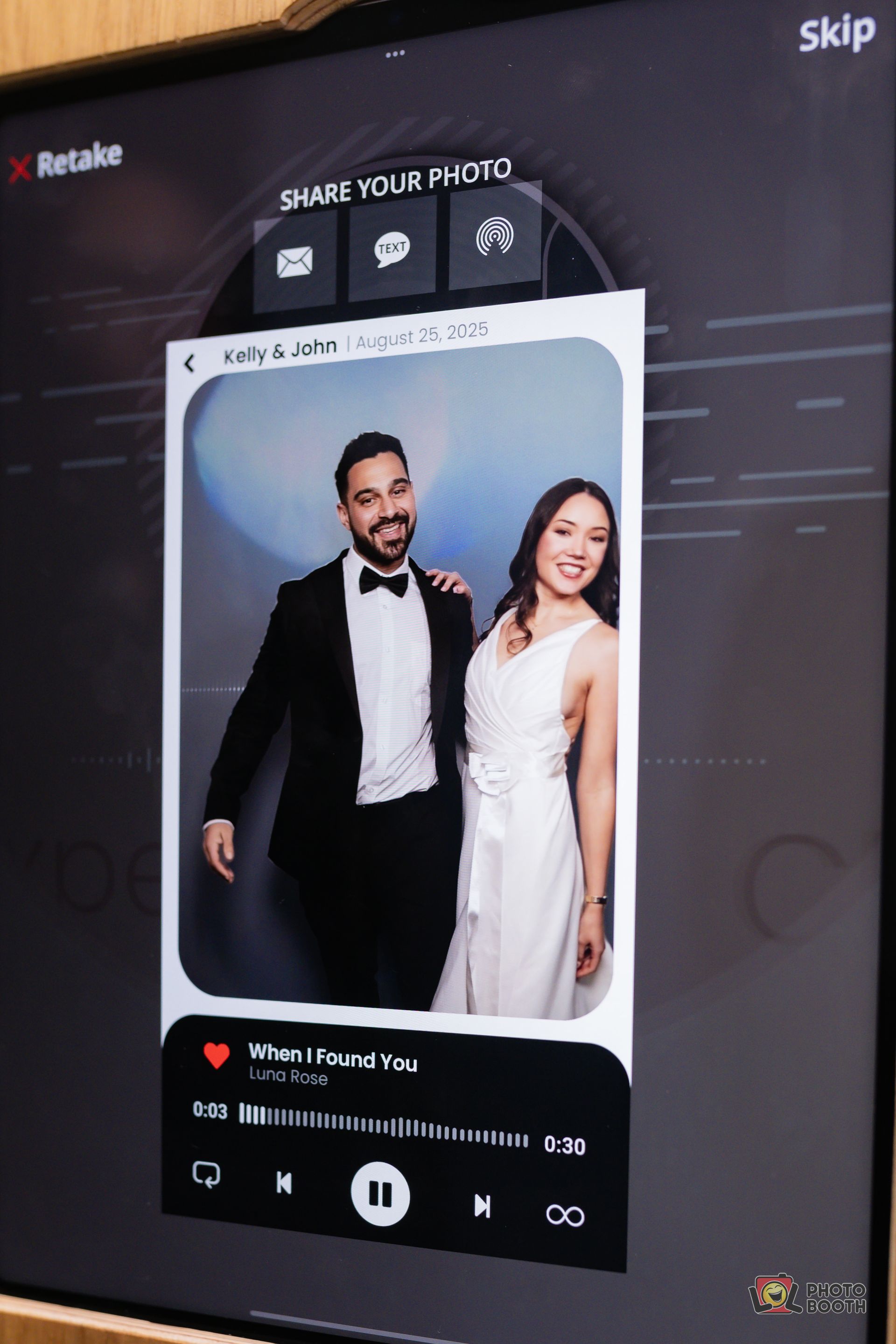 A formal photo booth image featuring a smiling couple, the man in a tuxedo and the woman in a white dress. They are against a white background.