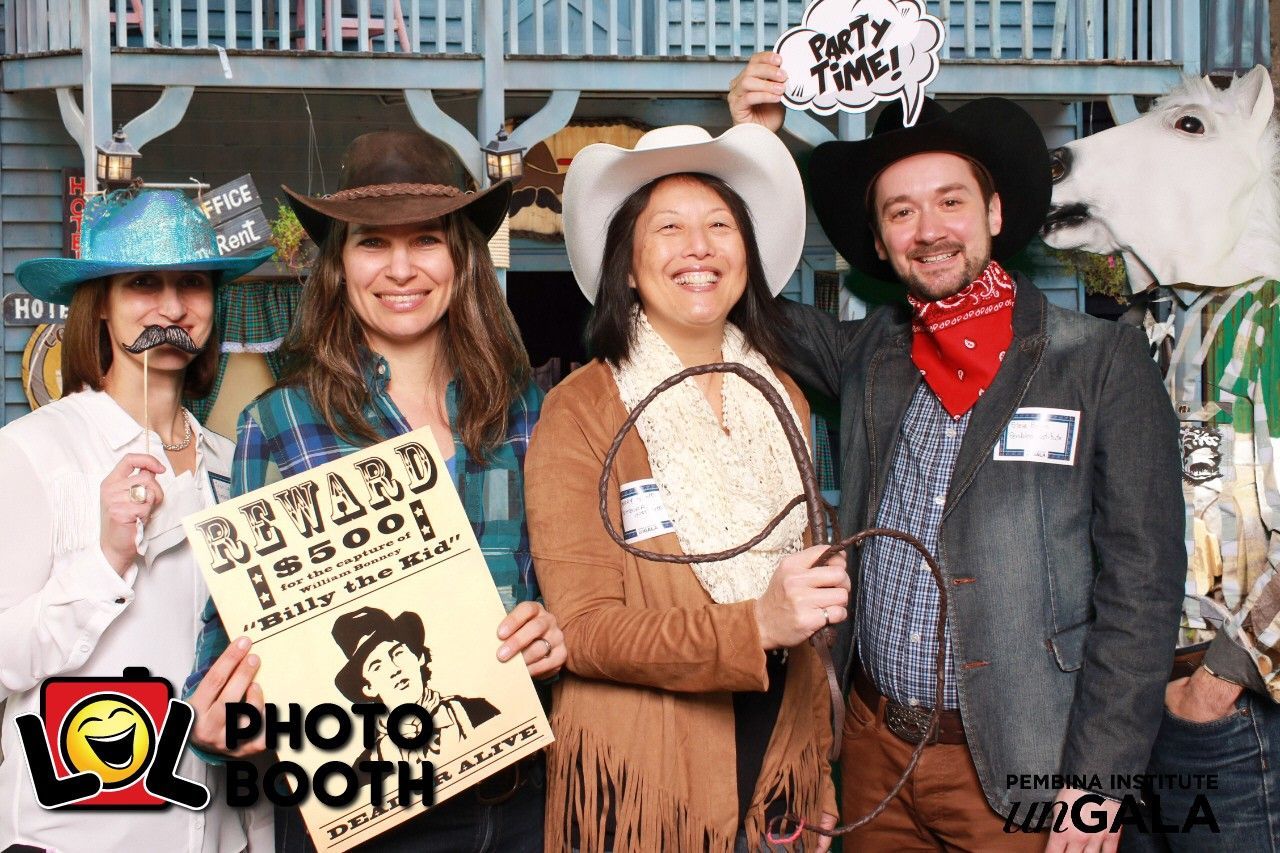 Four people pose for a photo booth in western costumes with props like a sign, hat, and lasso.
