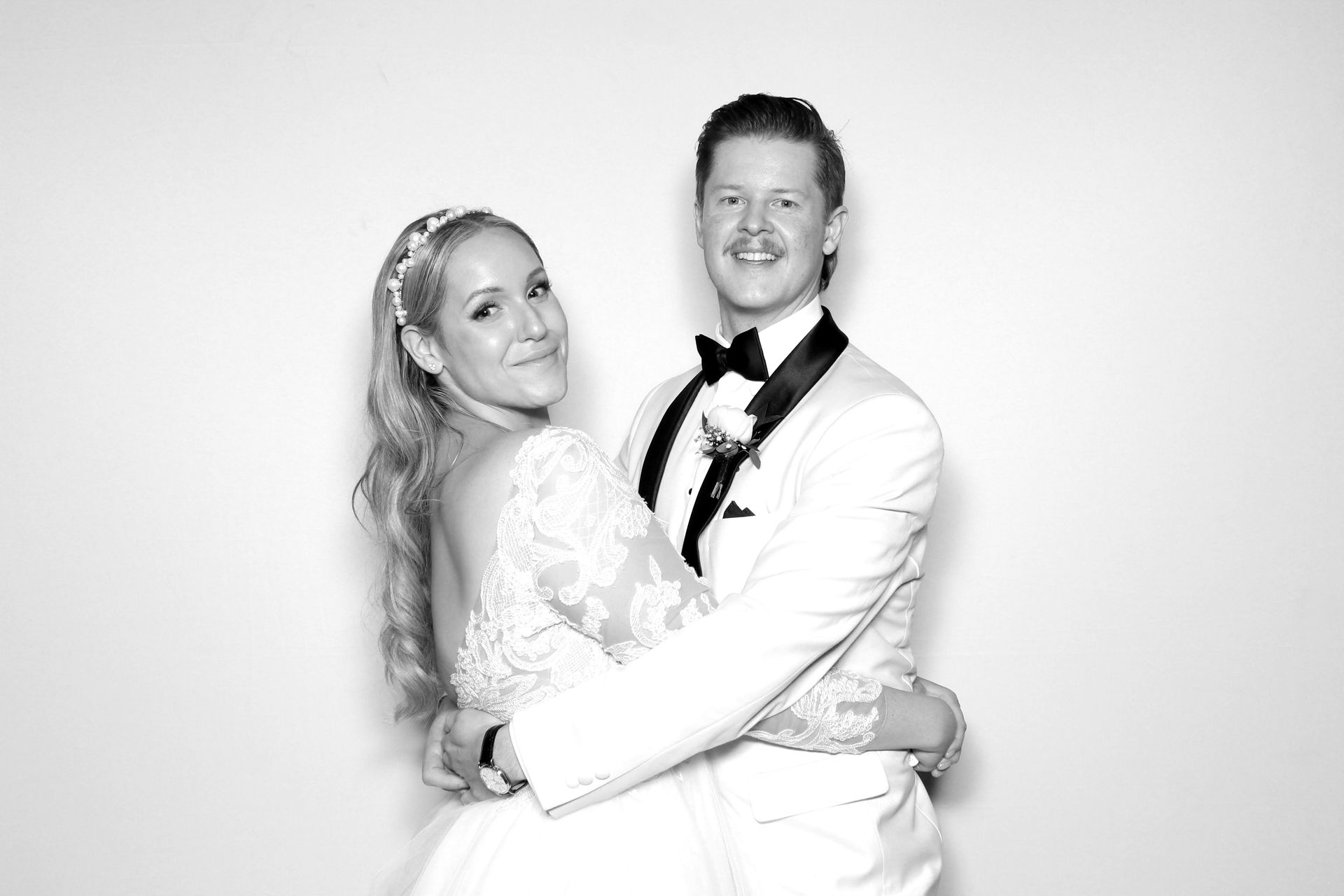 Newlyweds embrace, posing for photo booth. Bride in lace dress, groom in tuxedo. Black and white.