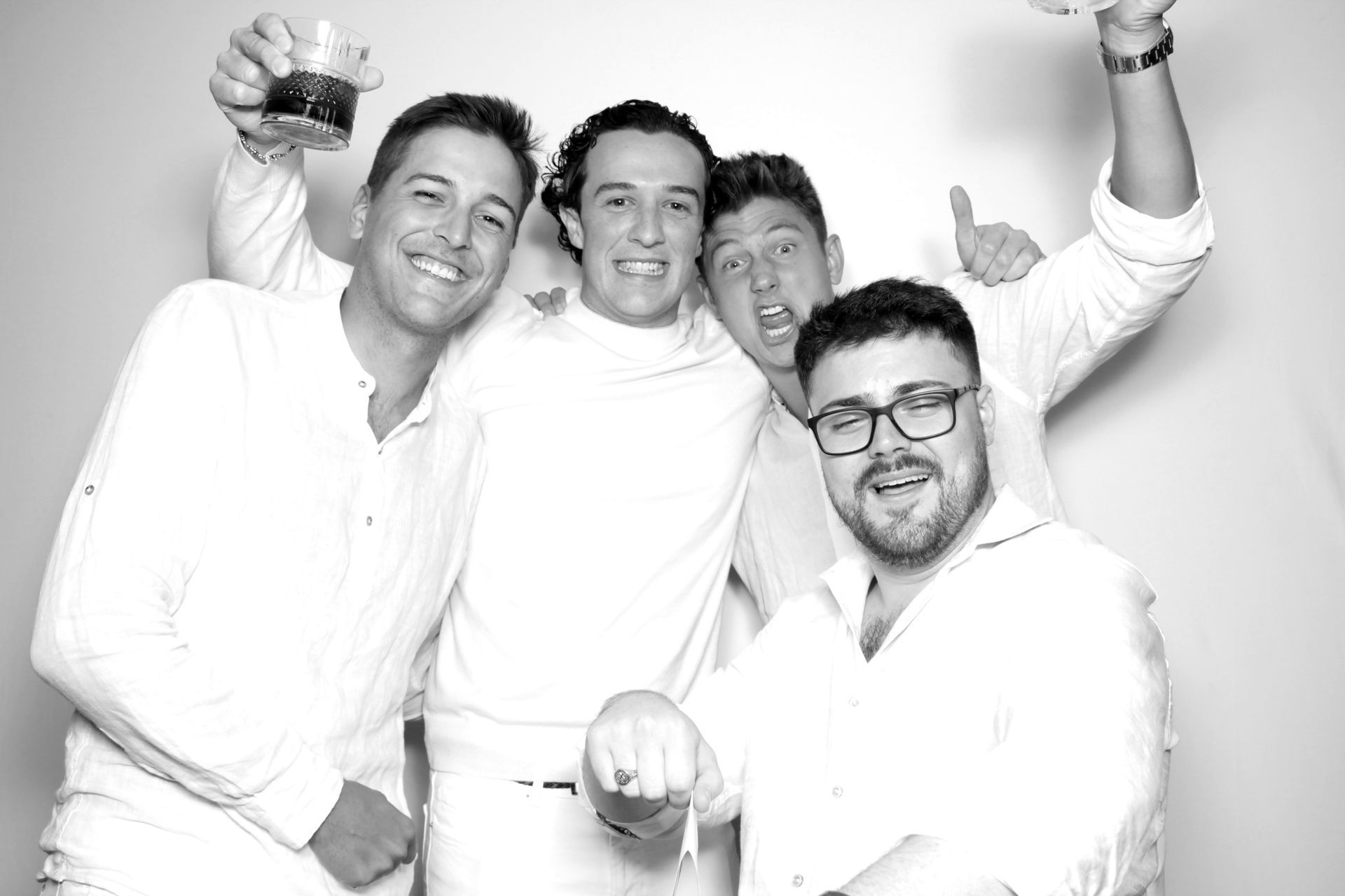 Four men in white shirts celebrating, holding drinks, and smiling in a photo booth.