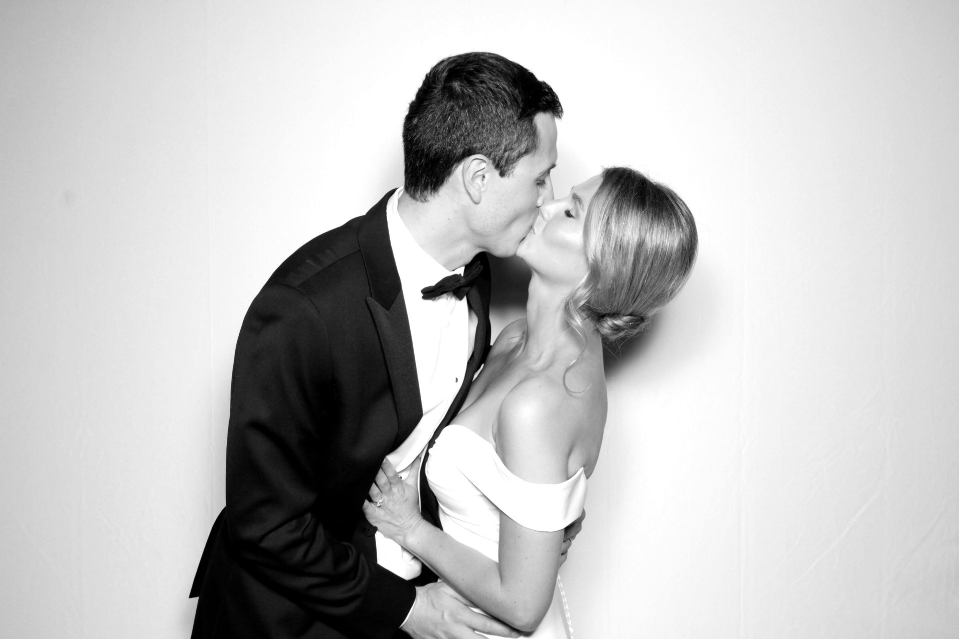 Couple kissing in a photo booth; the woman wears a white dress and the man a tuxedo.