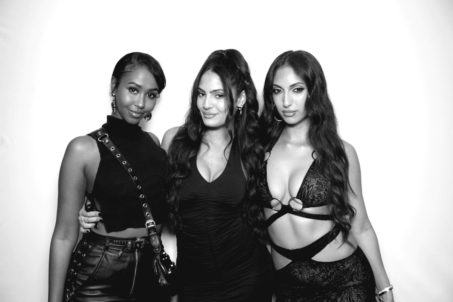 Three women pose together for a photo. They are in a black and white image and wearing fashionable outfits.