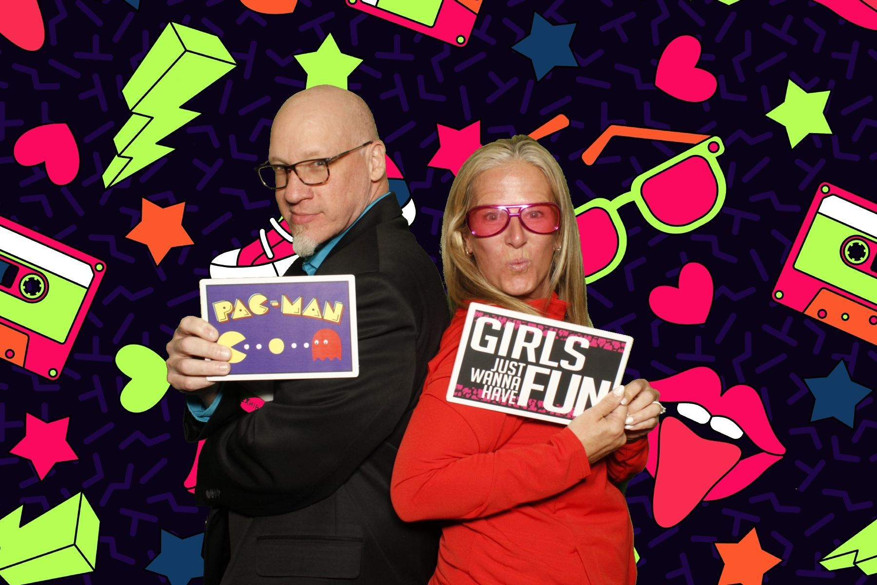 Two people posing with photo booth props against a retro background of neon shapes and cassette tapes.