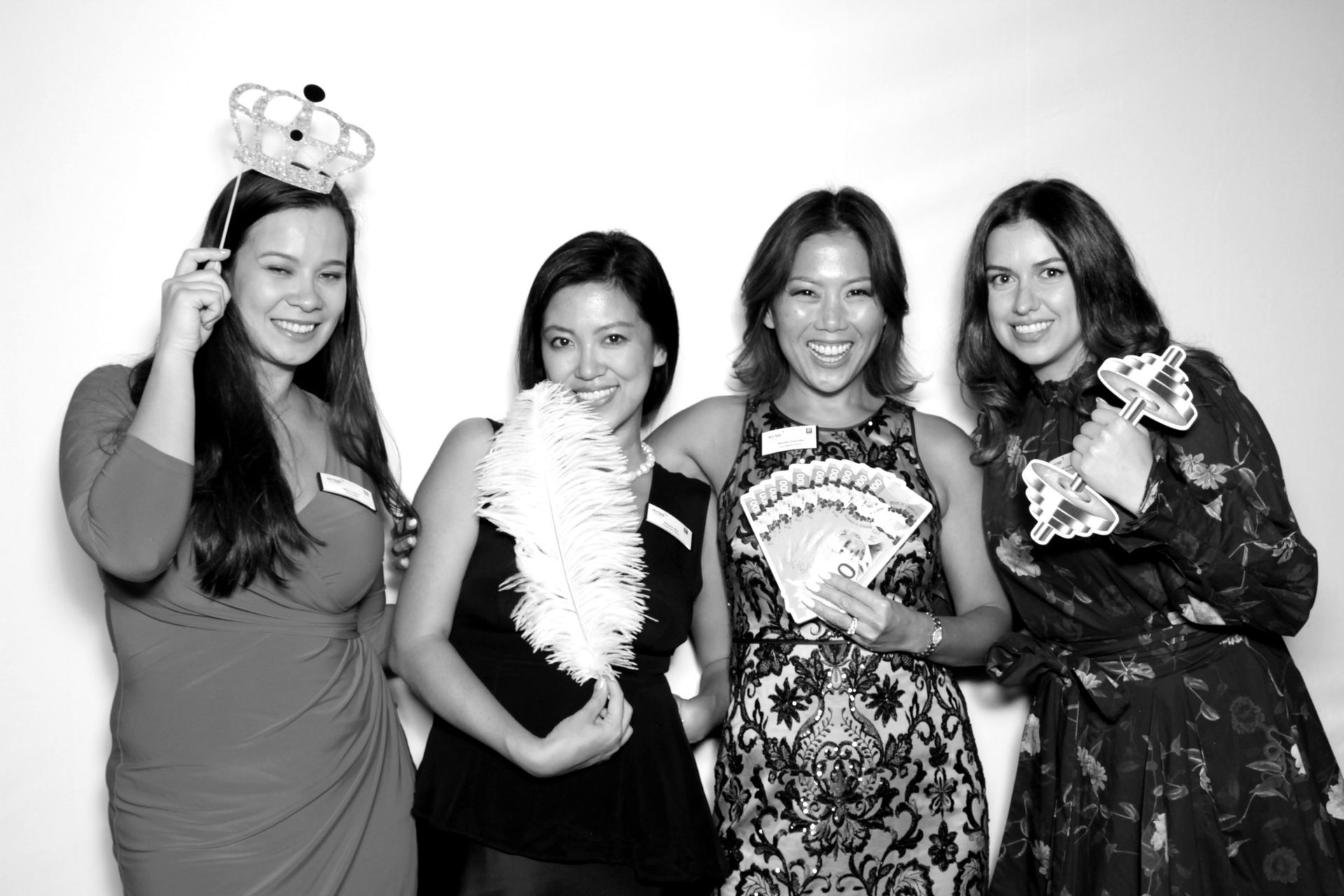 Four women posing with props in front of a white backdrop, smiling. One wears a crown, another holds a boa.