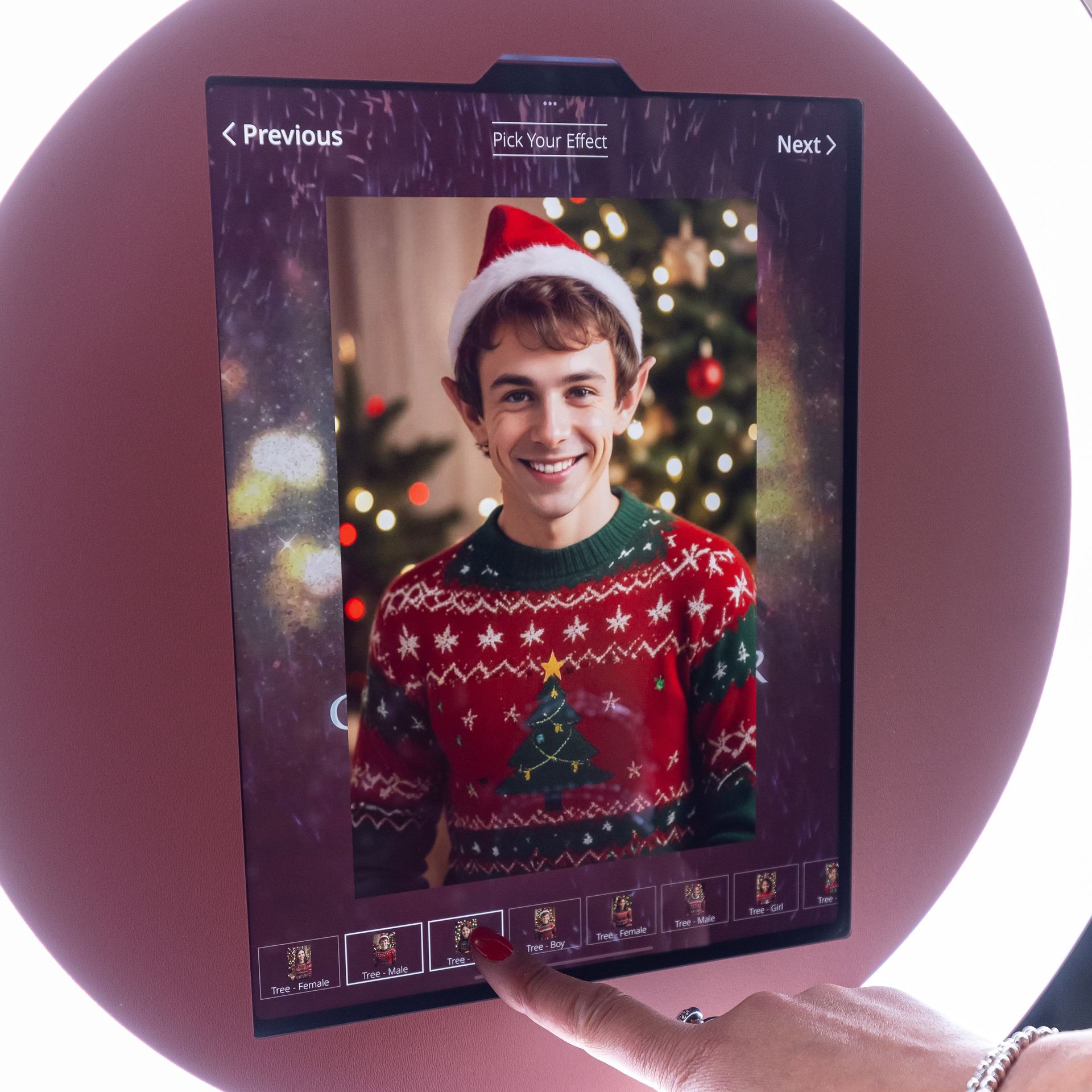 Man in Santa hat and Christmas sweater smiles at a photo booth screen, finger touching a selection.