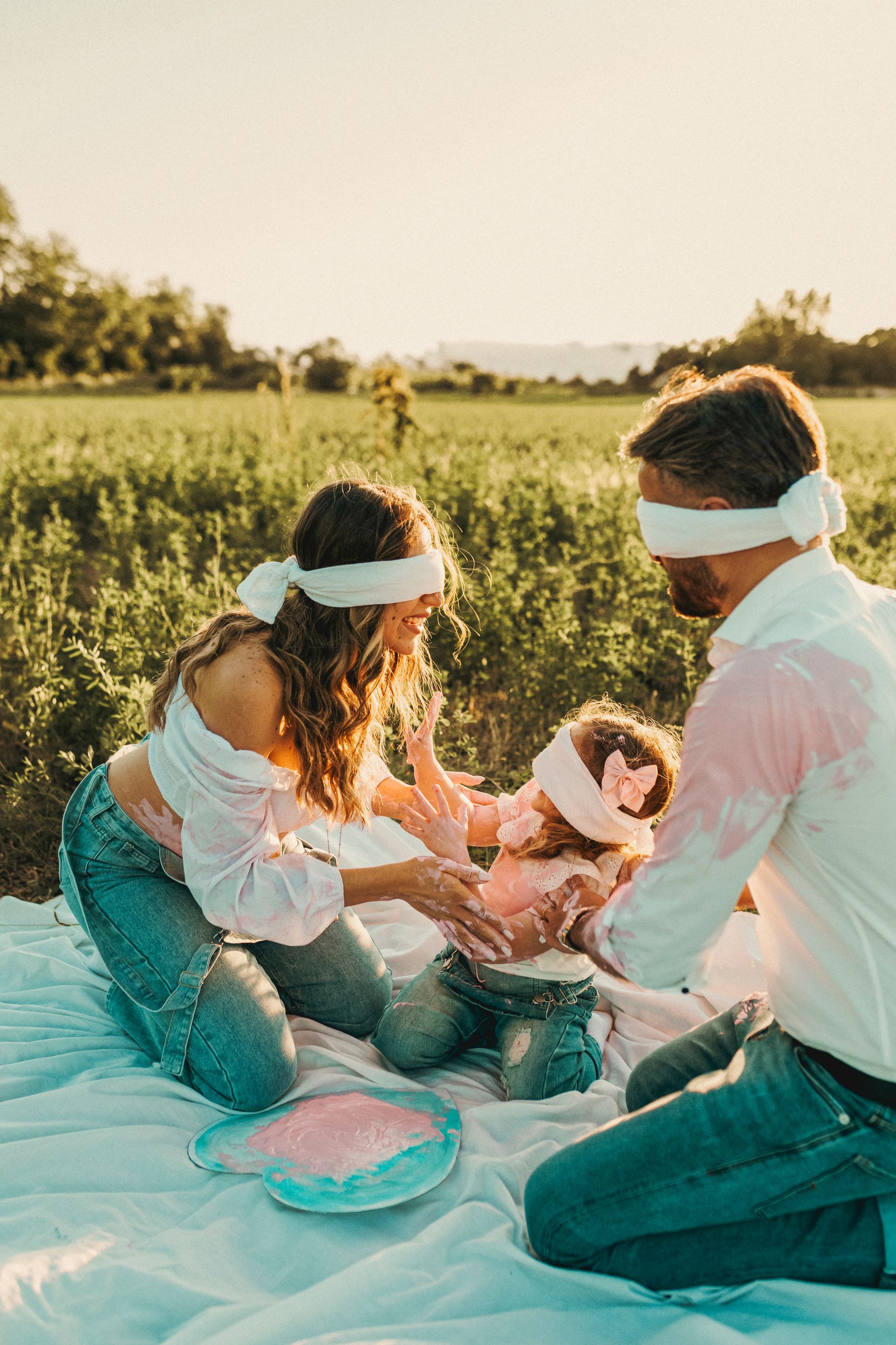 Famiglia, bendata, in procinto di rivelare il sesso del bambino. Cipria rosa sul padre. Ambientazione esterna, luce solare intensa.