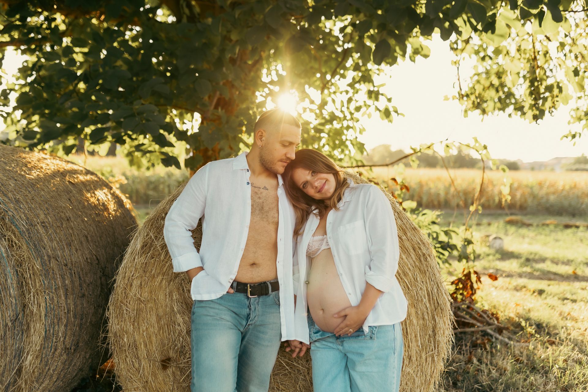 Coppia abbracciata vicino a balle di fieno in un campo, donna incinta, scena illuminata dal sole.