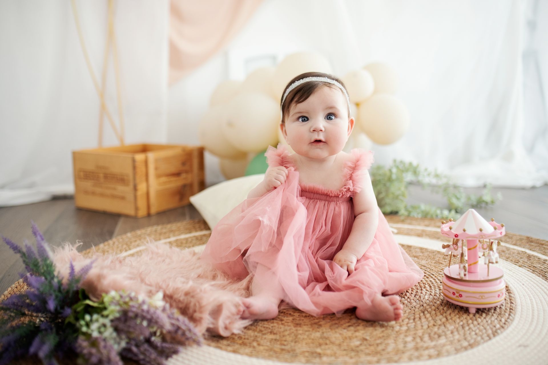 Un bambino con un vestito rosa, gli occhi spalancati, seduto su un tappeto rotondo decorato con palloncini e una giostra giocattolo.