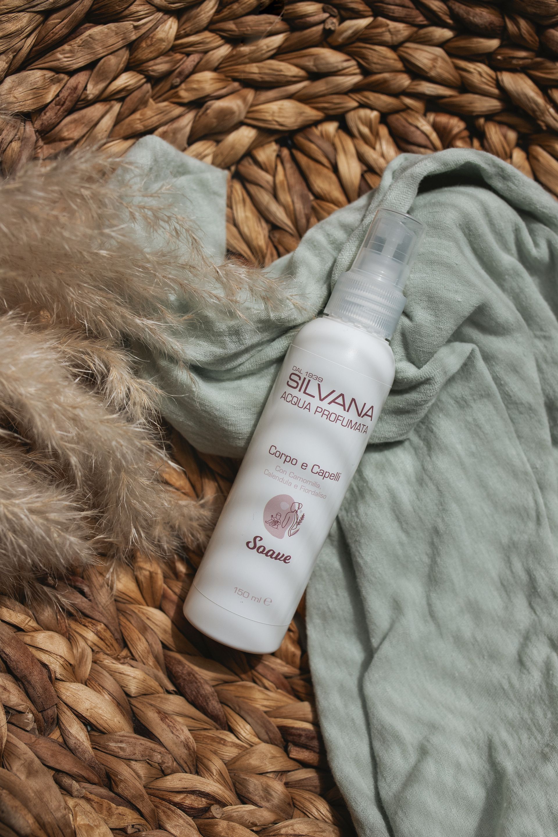 Flacone di prodotto per capelli Elvana su una superficie intrecciata con un panno verde drappeggiato e una pianta secca.