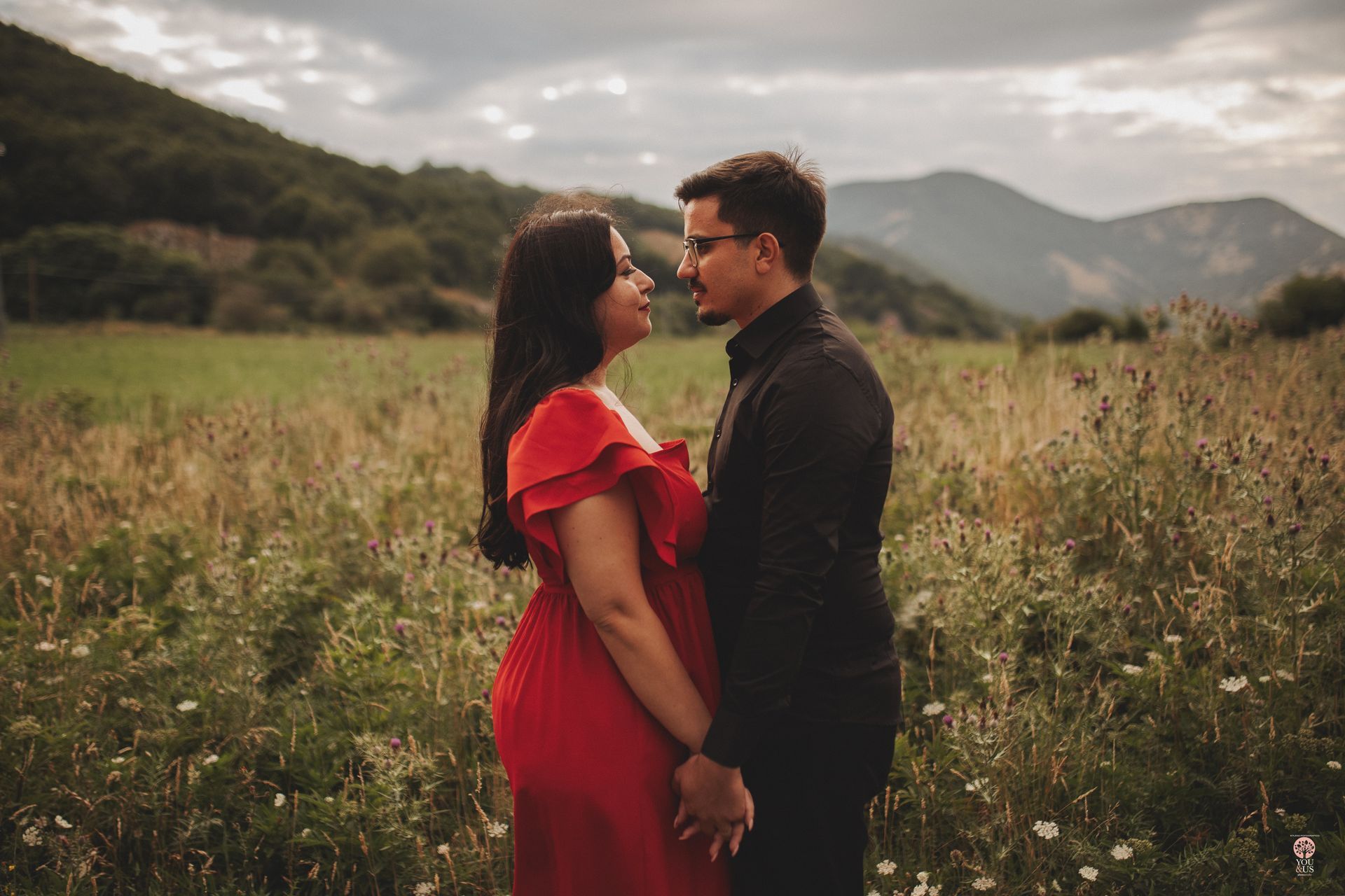Coppia che si tiene per mano, uno di fronte all'altro in un campo, donna in abito rosso, montagne sullo sfondo.
