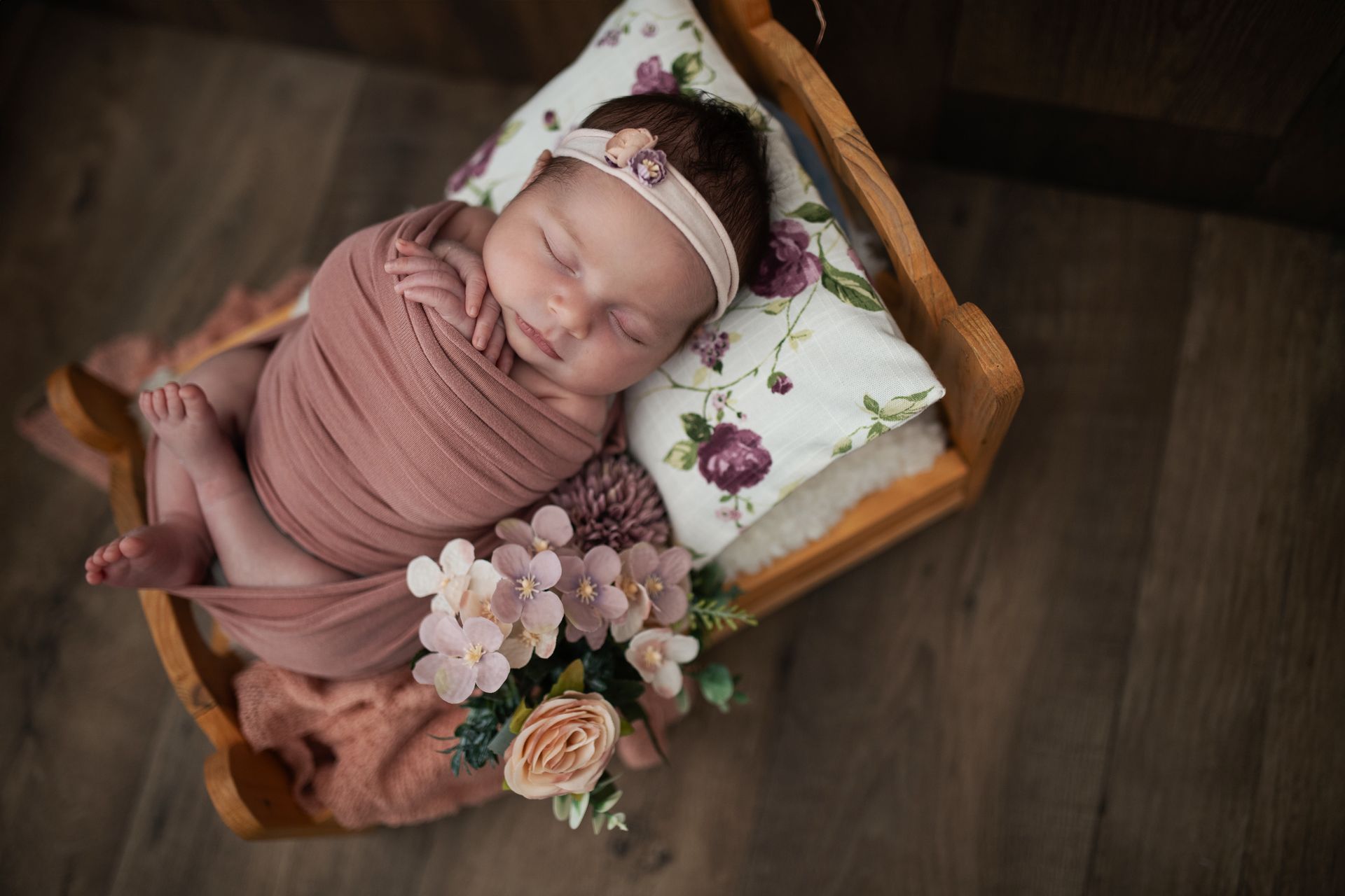Un neonato avvolto in una fascia rosa, dorme in un letto di legno decorato con fiori.