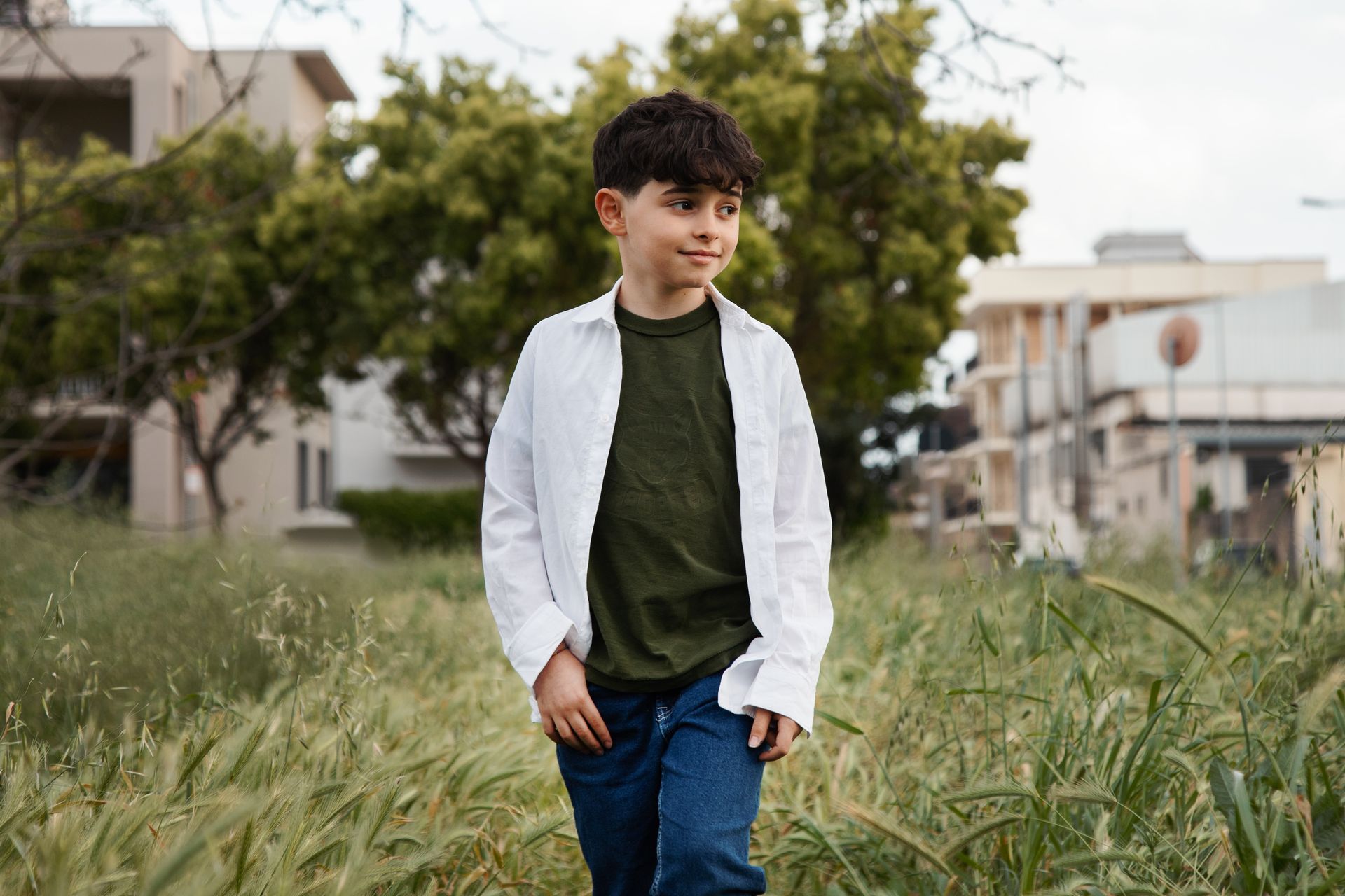 Un ragazzo cammina nell'erba alta, indossando una camicia bianca sopra una camicia verde e jeans blu, in un campo vicino agli edifici.