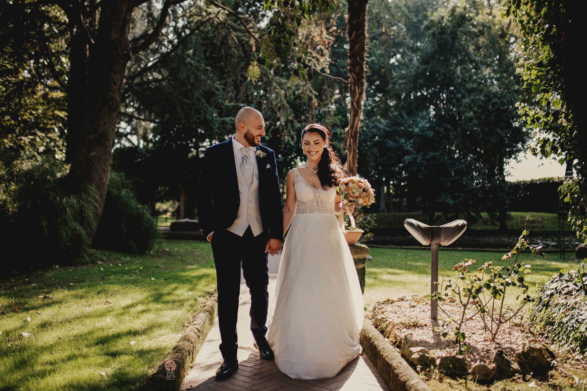 Due novelli sposi camminano mano nella mano in un giardino soleggiato. Lo sposo in abito elegante, la sposa in abito bianco, si sorridono.