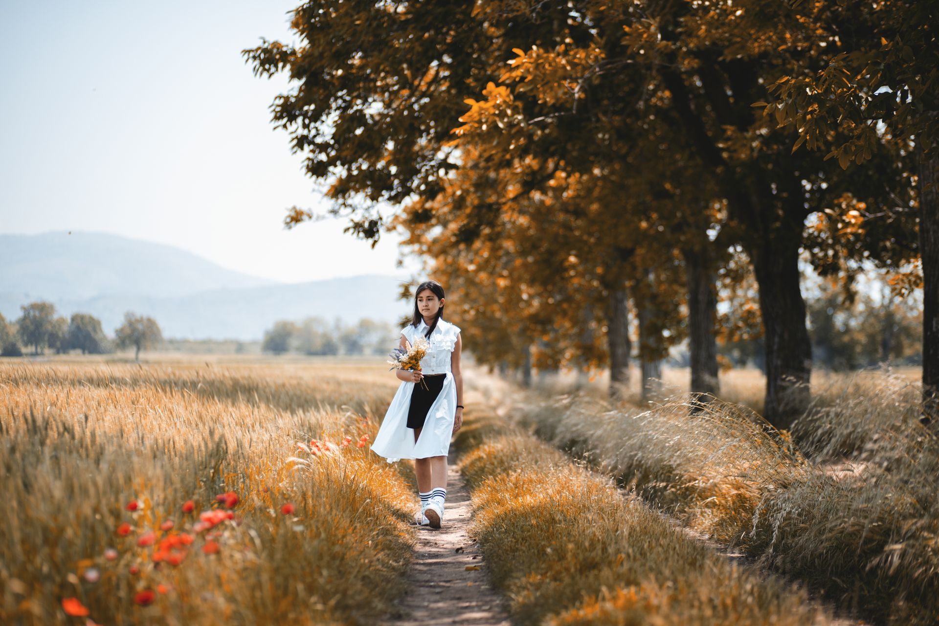 Una donna vestita di bianco cammina lungo un sentiero che costeggia un campo di grano e degli alberi, tenendo in mano dei fiori.